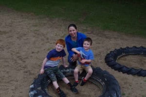 Woman and 2 young boys sitting on edge of large tire on playground