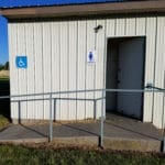 Small restroom building with "cowboys" sign at easy up tents and dog crates along side of agility ring at East Idaho Fairgrounds-BlackfootID