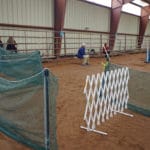 Agility ring entry gate with green construction mesh fence and white baby-gate opening at Green mesh construction fencing to make chutes out of agility ring with white folding baby gate on entrance side at South St. Louis County Fairgrounds, Dirt Floor Arena, Proctor MN